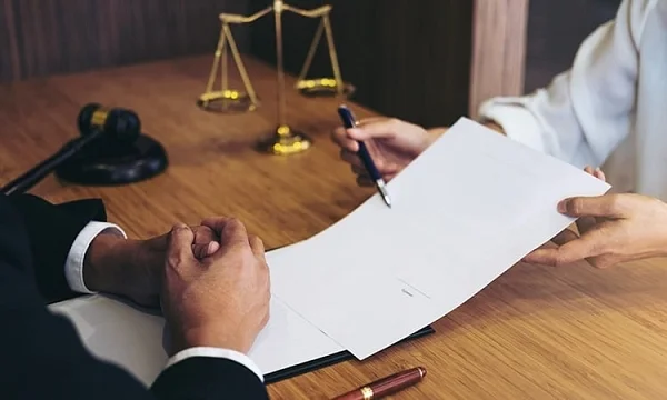 Person handing a document and pen across a desk, with scales of justice and a gavel behind